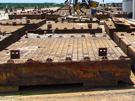 Medium-format photograph: Pad Deck, Launch Complex 39-B, east side of Flame Trench. Removed Crawlerway Grid Panels lie stacked on cribbing. Note the temporary lifting lugs (edge-up, with 2 holes in each one of them for the attachment of lifting hardware) which were welded to the top flanges of the Mystery I-Beams on every Panel, and which were used for Panel lifting operations because the original Ring Bars recessed into the Panel surfaces were corroded beyond any useful degree of trustworthiness and could not be used. Photo credit: Withheld by request. Medium-format photograph: Pad Deck, Launch Complex 39-B, east side of Flame Trench. Removed Crawlerway Grid Panels lie stacked on cribbing. Note the temporary lifting lugs (edge-up, with 2 holes in each one of them for the attachment of lifting hardware) which were welded to the top flanges of the Mystery I-Beams on every Panel, and which were used for Panel lifting operations because the original Ring Bars recessed into the Panel surfaces were corroded beyond any useful degree of trustworthiness and could not be used. Photo credit: Withheld by request.
